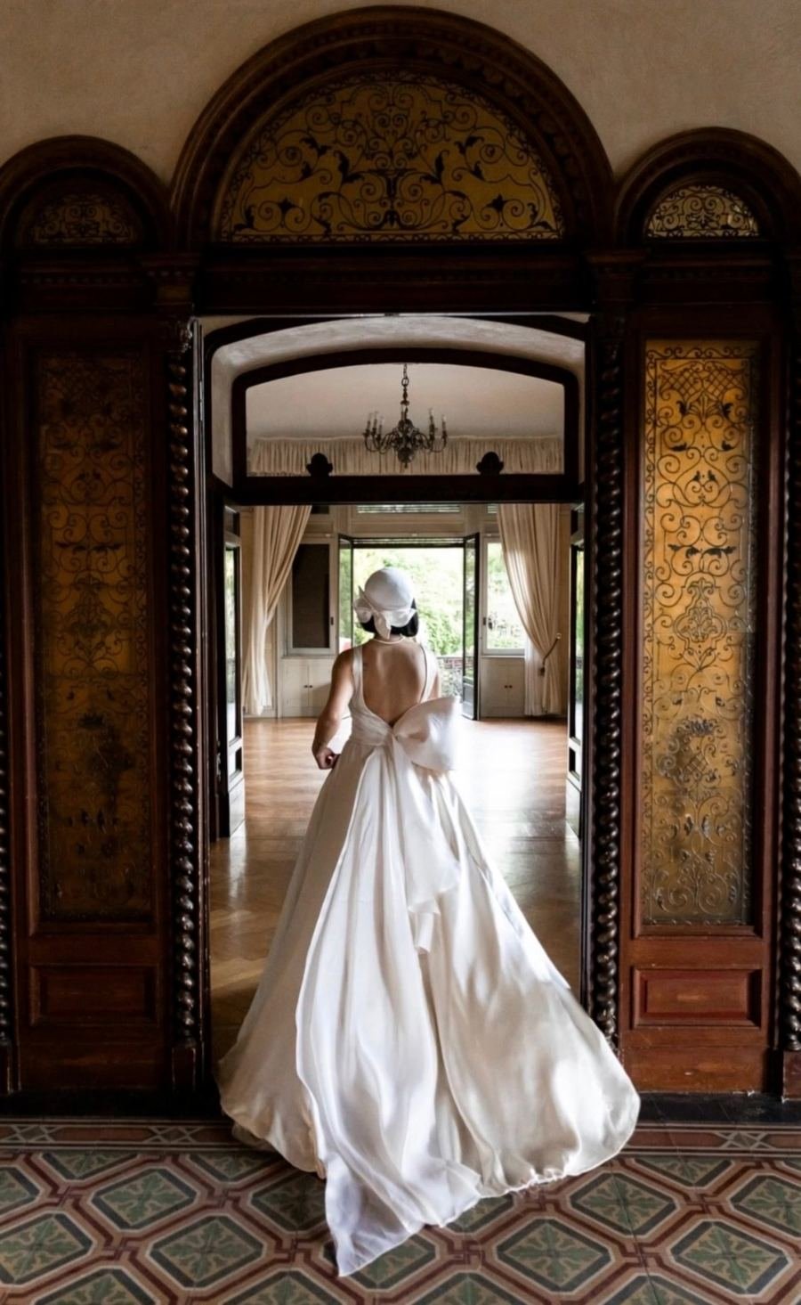 Una sposa Maison Bourelly, di spalle, attraversa le sale di una villa con l’abito in raso dal grande fiocco e il cappello coordinato